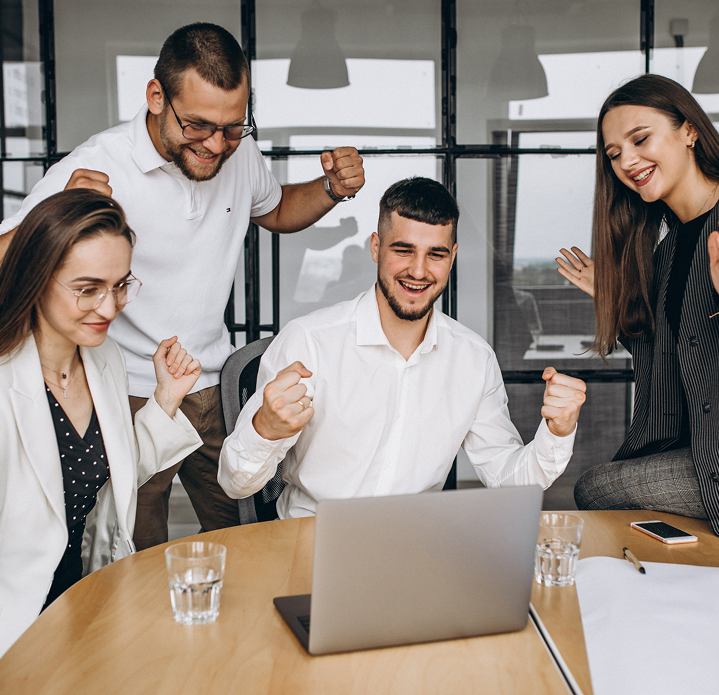 Group of four professionals celebrating success around a laptop in a modern office setting.