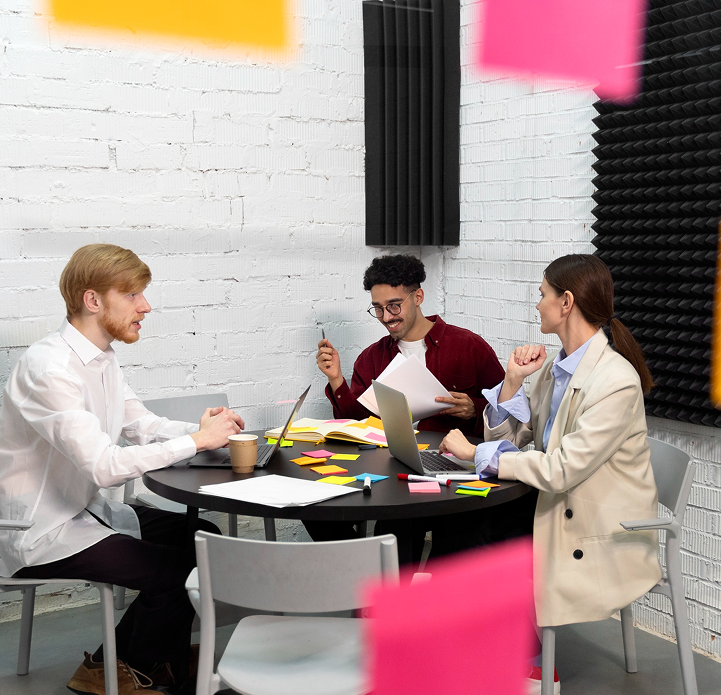 Three professionals collaborate at a round table, surrounded by colorful sticky notes and laptops in a modern office setting.
