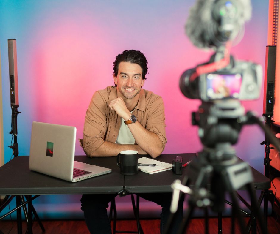 Young man smiling at a desk with a laptop and coffee cup, set against a colorful backdrop, ready for a video shoot.