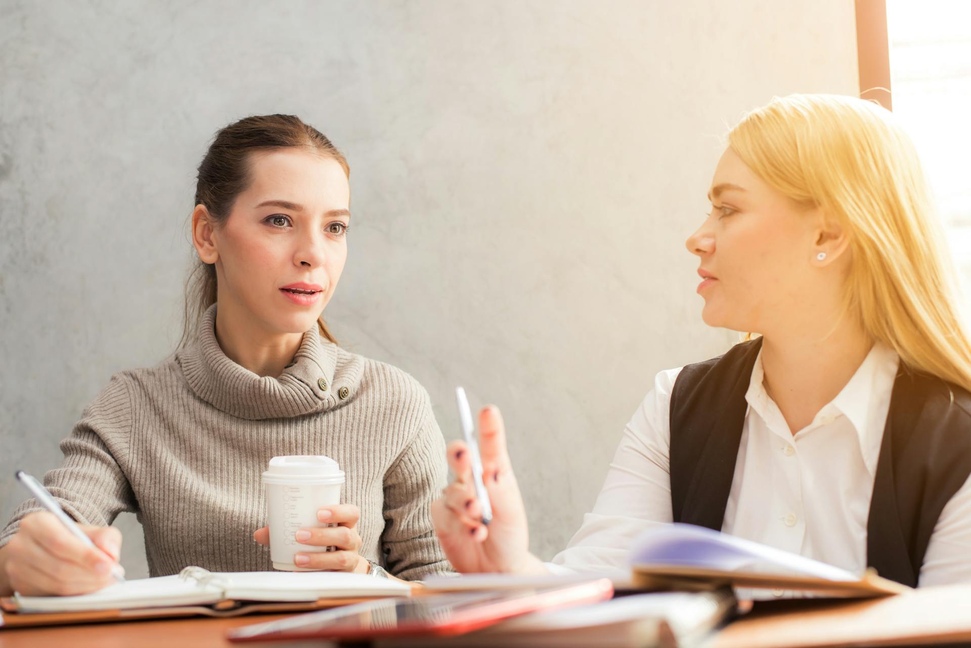 Two women engaged in a discussion at a table, one holding a coffee cup and the other gesturing with a pen.