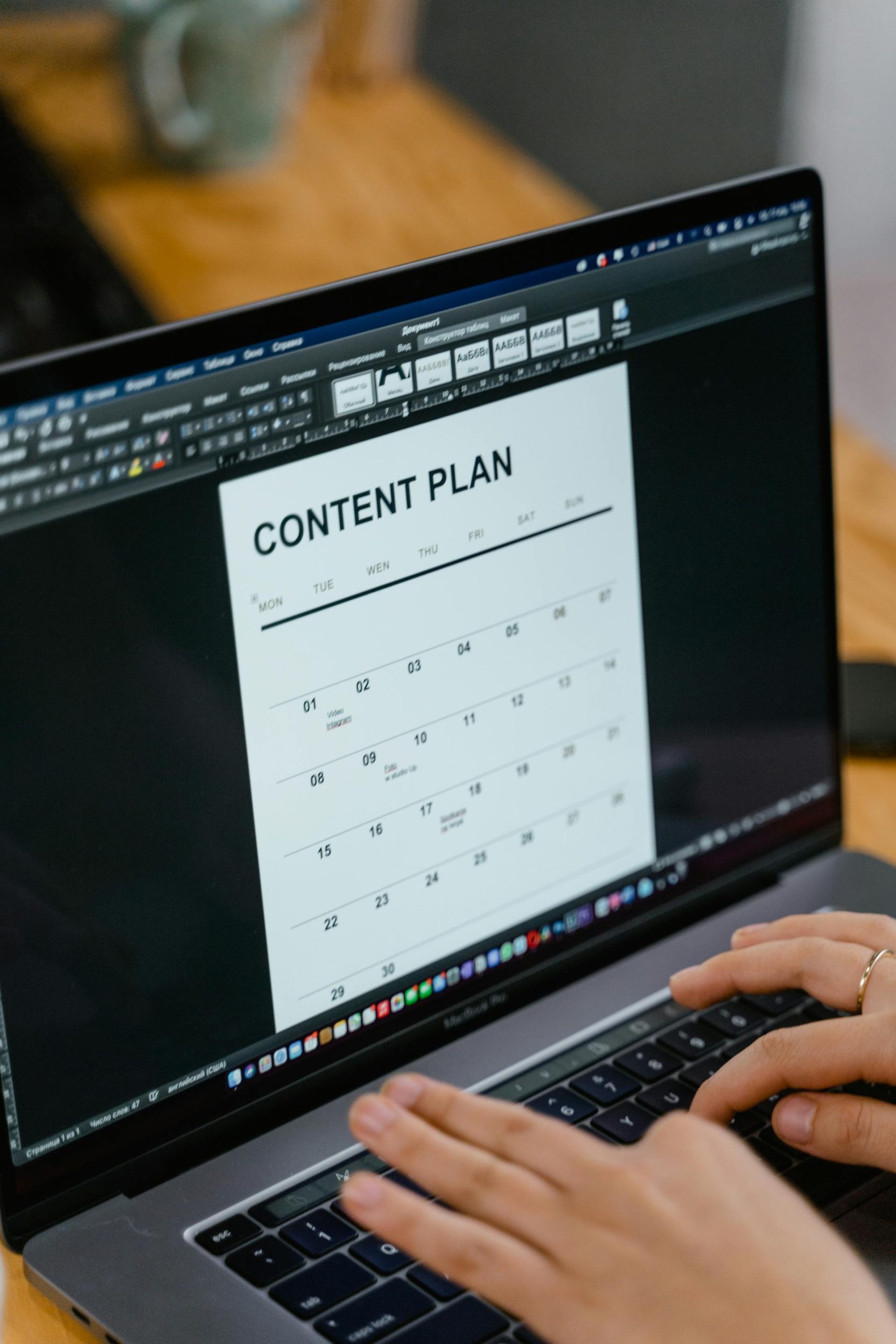 Person typing on a laptop displaying a content plan calendar on the screen, set on a wooden desk.